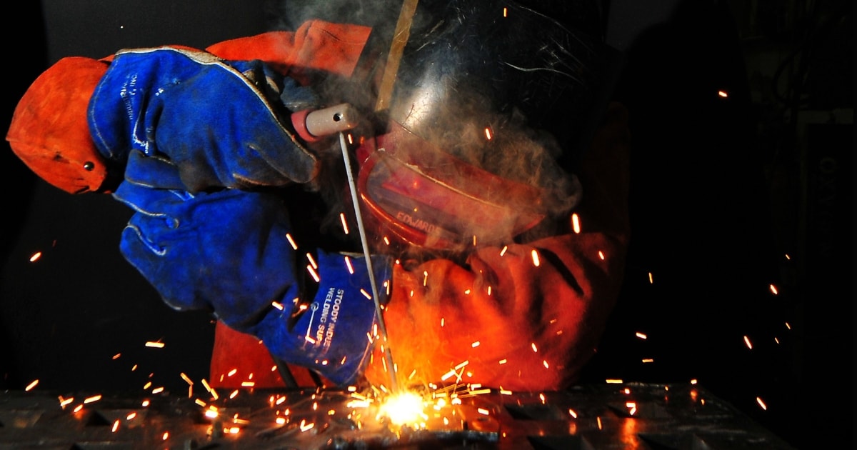 Welder wearing full protection gear including helmet, suit, and gloves while welding metal.